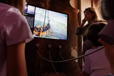 L'image montre un groupe de personnes, dont une femme et des enfants, observant un écran. L'écran présente une image d'un bateau sur l'eau, probablement dans un contexte historique ou éducatif. Les participants semblent écouter des informations à travers des casques audio reliés à l'écran. L'environnement est chaleureux, et les murs sont décorés de manière rustique, ce qui ajoute à l'atmosphère immersive de l'expérience.