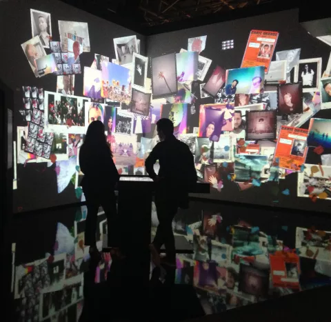 L'image montre une installation artistique immersive, où des projections de diverses photos et affiches couvrent les murs et le sol d'un espace sombre. Deux silhouettes de personnes se tiennent devant cette exposition, observant les images qui les entourent. L'ambiance semble à la fois moderne et artistique, avec une variété d'images qui évoquent différentes émotions et souvenirs.