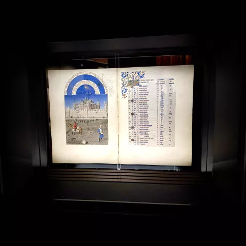 L'image montre un ancien manuscrit exposé dans une vitrine. Sur la gauche, une illustration colorée représente une scène avec des personnages, probablement à l'extérieur, devant un bâtiment imposant qui pourrait être un palais ou une cathédrale. Sur la droite, on trouve une liste écrite, probablement des noms ou des événements, ornée d'initiales décoratives en couleur. Le fond est sombre, mettant en valeur le manuscrit et ses détails complexes.