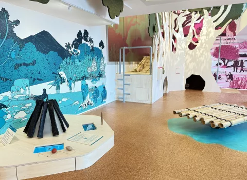 L'image montre un espace d'exposition, probablement dans un musée ou un centre éducatif. On y voit des murs décorés de peintures représentant des paysages et des scènes de la nature. À gauche, il y a une structure en forme de pyramide, faite de matériaux sombres, qui semble être interactive. Au centre, il y a une série de barres en bois disposées à l'horizontale, rappelant un radeau. Le sol est recouvert d'un matériau de couleur claire, et il y a des éléments d'eau stylisés sur le sol. L'ensemble de l'espace semble être dédié à l'exploration de la nature et de l'environnement, avec une approche ludique et éducative.