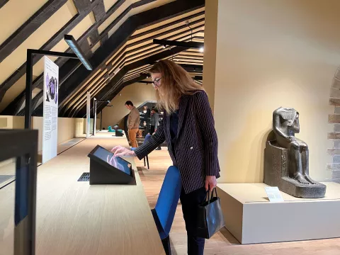 L'image montre une femme se tenant près d'un comptoir dans un musée. Elle semble interagir avec un écran tactile. En arrière-plan, on peut apercevoir d'autres visiteurs et une statue ou une sculpture ancienne sur un socle. La pièce a des poutres en bois au plafond, créant une ambiance chaleureuse et accueillante. La femme porte une tenue professionnelle avec un sac à main.