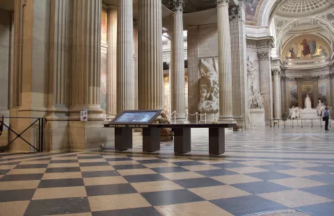 L'image montre l'intérieur d'un grand bâtiment avec des colonnes majestueuses et un sol en damier. On peut voir des sculptures et des fresques sur les murs, ajoutant à l'impression d'élégance et d'histoire de l'espace. Au centre, il y a une table interactive qui semble fournir des informations aux visiteurs. L'éclairage est doux, mettant en valeur les détails architecturaux. Des personnes se déplacent, ce qui indique que c'est un lieu ouvert au public.
