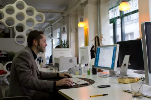 The image depicts a man sitting at a desk in an office environment. He is focused on working on a computer, which has a blue screen displaying a document or application. The office space appears modern and open, featuring large windows that let in natural light. There are several other computers and office items in the background, along with some plants that add a touch of greenery to the workspace. The man is dressed in a brown coat and has a look of concentration as he engages with his work.